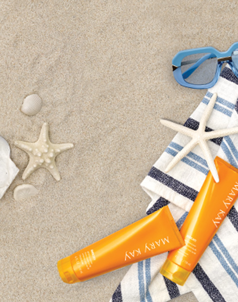 Two bottles of Mary Kay Sun Care Sunscreen styled against sand, striped beach towel and beach accessories.