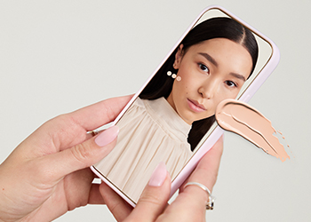 Light-skinned hands holding phone showing scan analysis of woman’ skin, with light foundation shade swipe, on gray background