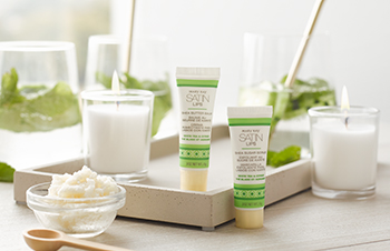 Satin Lips Set in green and white tubes with light yellow caps on a countertop with shea butter and candles