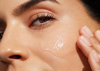 Up close macro image of a brunette woman confidently looking into the camera while applying skin care to her cheek
