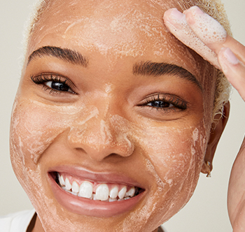 A smiling black woman exfoliating with the Botanical Effects Invigorating Scrub.