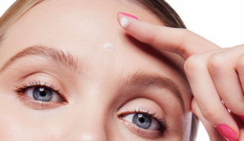 Blonde woman wearing hot pink nail polish rubbing in Dynamic Wrinkle Limiter topical cream to targeted spots on her forehead
