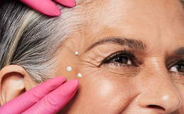  pink medical gloved hand rubbing in Dynamic Wrinkle Limiter topical cream to a grey-haired woman’s extended crow’s-feet area