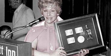 Mary Kay Ash smiles for a photo while standing at a podium and holding up a plaque.