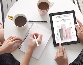 Two women are working at a table with coffee cups, a notebook, and an iPad that shows an image of the new TimeWise Miracle Set 3D from Mary Kay.