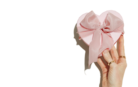 Baby Pink heart-shaped box with a pink ribbon and bow being held by a light-skinned hand with rings on, on a plain white background